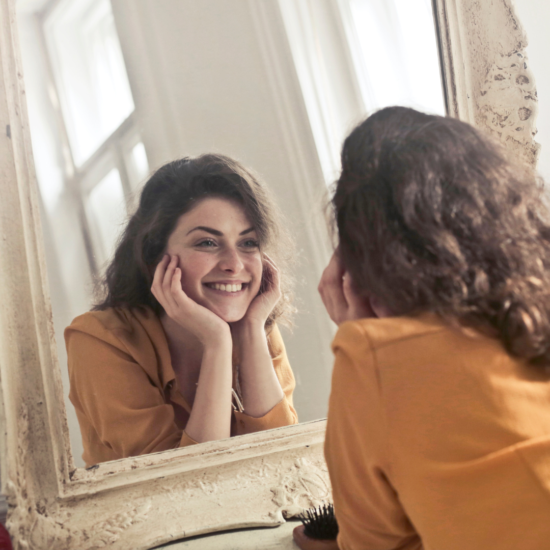 Mujer sonriendo ante el espejo, con las manos apoyadas en las mejillas.